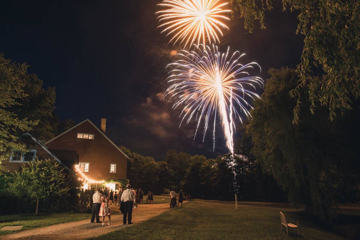 Night scene of colorful fireworks bursting in the sky above a small illuminated house surrounded by trees and people watching the fireworks display at stonover farm in the Berkshires