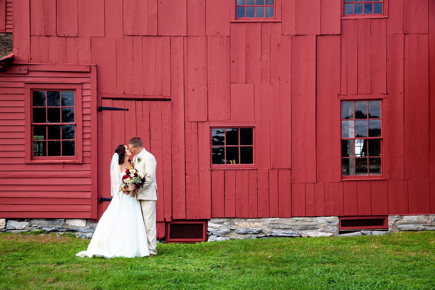 The bride and groom sharing a kiss in front of a red barn at hancock shaker village