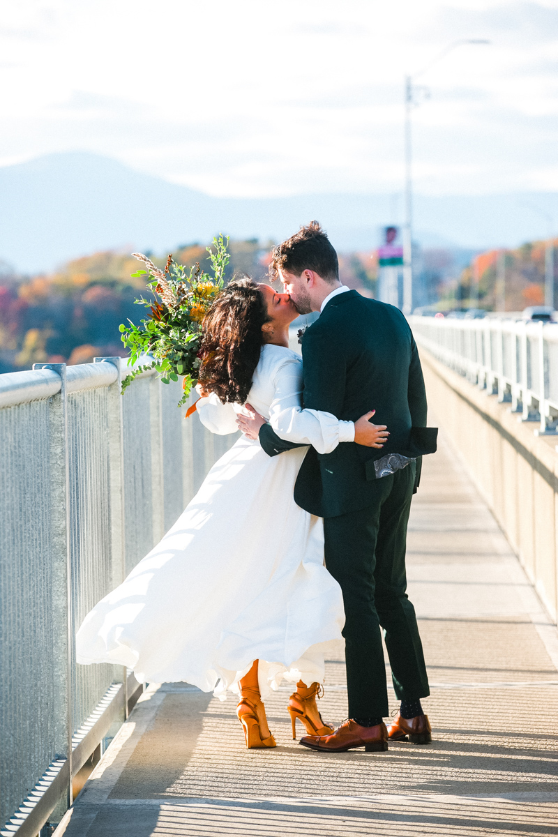 Bride and groom sharing a kiss on a bridge with scenic fall mountains in the background, the bride holds a bouquet, and the groom in a dark green suit embraces her, autumnal sunlight highlights the scene.