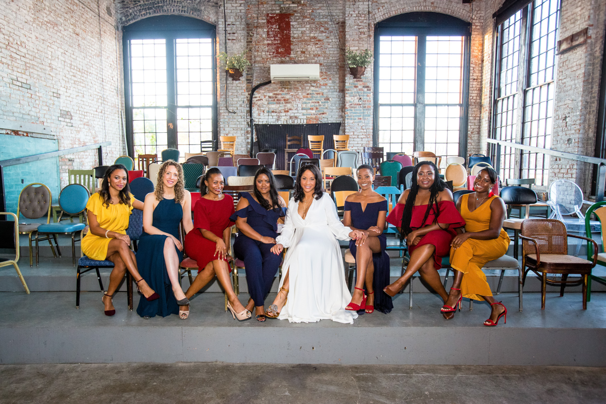 Bride in a flowing white dress seated center among bridesmaids in colorful dresses inside an industrial-chic venue with exposed brick, large windows, and assorted chairs at Basilica Hudson.