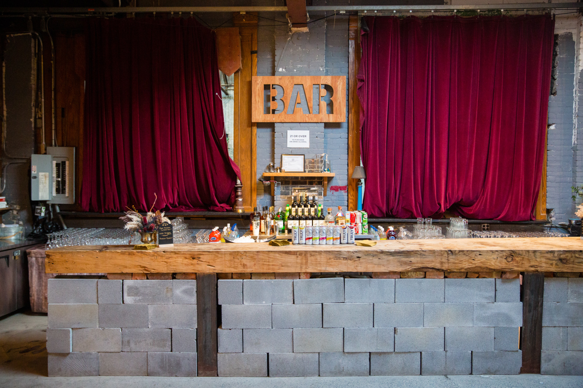 The image shows a rustic wedding bar setup in an industrial venue with a backdrop of maroon curtains and exposed brick walls. The bar itself is made of a live-edge wooden countertop on cinder block legs, adorned with an array of bottles, glassware, and a small floral arrangement, giving a warm and inviting atmosphere for the guests.