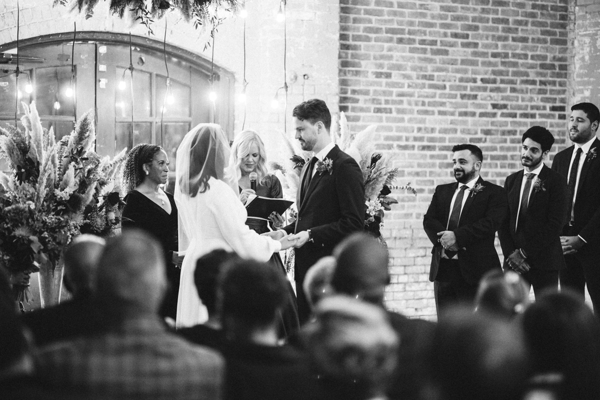 The image is a black and white photo capturing an intimate moment during a wedding ceremony at Basilica Hudson. The couple is at the altar, exchanging vows with the officiant in the background, surrounded by tall, wildflower arrangements. The groom, in a sharp suit, faces the bride, who is wearing a veil, with an onlooker's perspective from behind the seated guests. The industrial character of the venue, with its brick walls and large windows, creates a striking contrast against the soft, romantic moment of the couple.