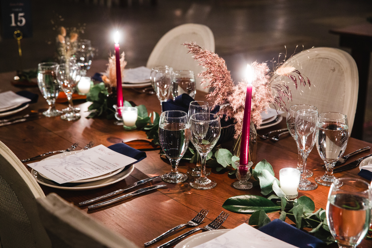 The image showcases a close-up view of a wedding reception table at Basilica Hudson, intimately lit by candlelight. The rustic wooden table is adorned with elegant crystal glassware, silver cutlery, and vibrant pink candles among greenery and fluffy pampas grass, creating a warm and sophisticated atmosphere.