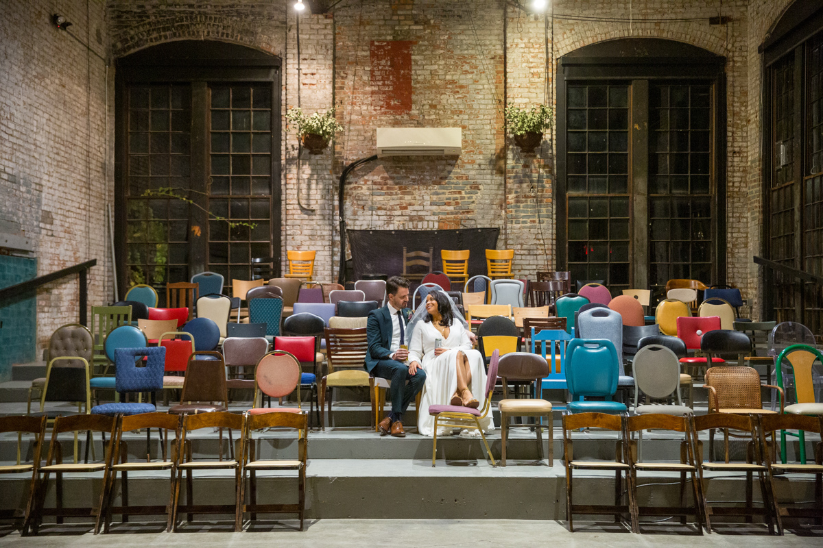 A bride and groom are seated together on a stage in an empty hall at Basilica Hudson, surrounded by a colorful array of mismatched chairs. The room's vintage charm is highlighted by the exposed brick and hanging greenery, creating a warm, intimate atmosphere.