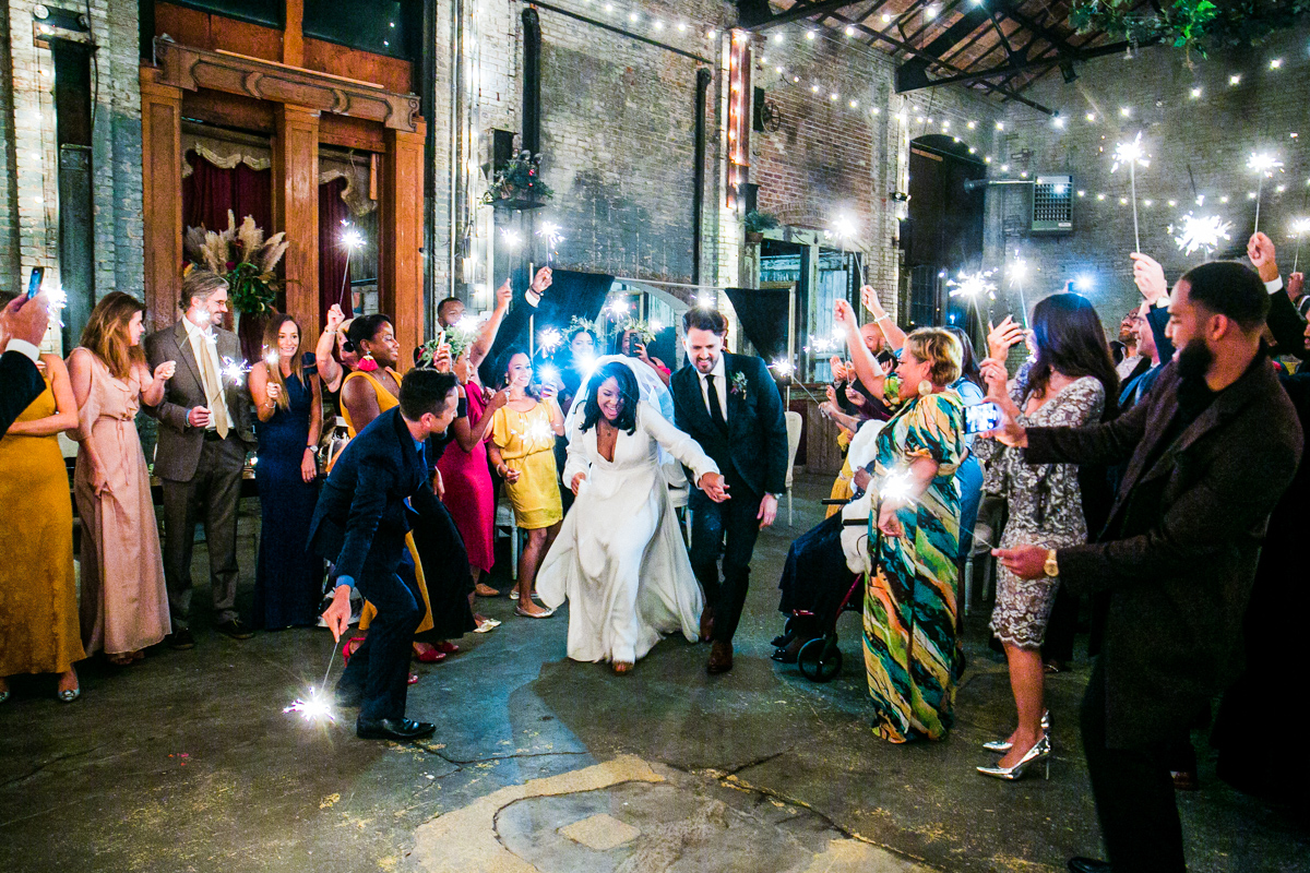 The image shows a bride and groom making a joyful entrance through a sparkler welcome, with guests lining the path, celebrating the moment. The industrial space of Basilica Hudson is filled with the warm light of the sparklers, adding to the festive ambiance.