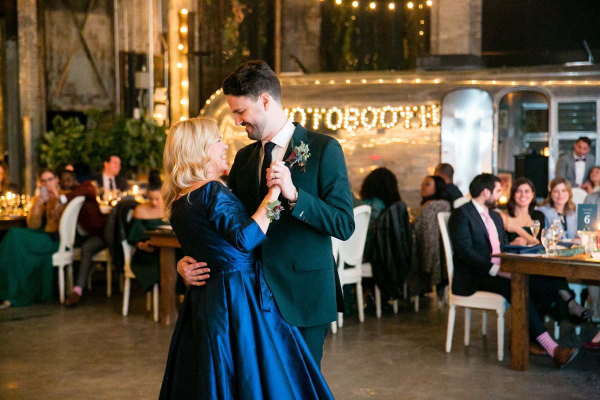 A tender moment is captured as the groom, dressed in a dark green suit, shares a dance with his mother in a deep blue dress. They are both smiling, enjoying this special moment, with the festively lit photo booth and seated guests in the background at Basilica Hudson.