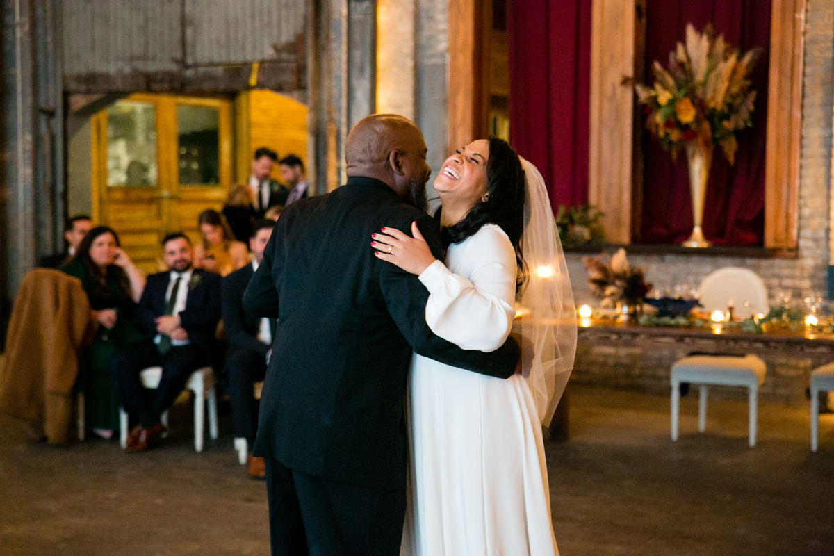 The bride, radiant in her white dress and veil, shares a dance with her father, whose expression is filled with affection and pride. They're surrounded by the warm ambiance of the Basilica Hudson venue.