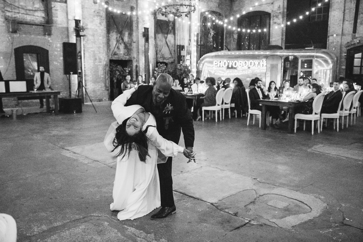 
In this black and white image, a bride shares a dance with her father during her wedding at Basilica Hudson. They are captured in a playful moment, with her father leading her in a dramatic dance move that showcases their joy and the celebratory spirit of the occasion.